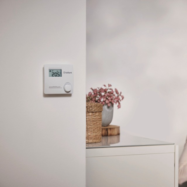 a sensoROOM pure heating control on a wall, a woman is sat on the sofa in the background with a mug in her hand.