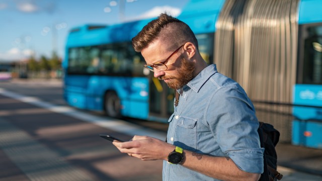 man outside looking at his phone while a bus is driving behind him