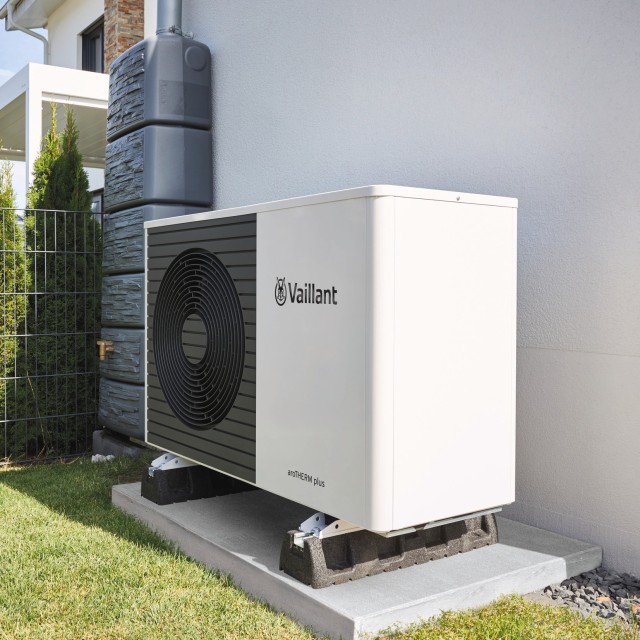 A aroTHERM plus heat pump facing towards the left hand side. The unit is places against a wall with grass and tress 